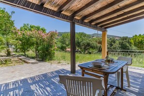 Eine Terrasse im Freien mit Tisch und Stühlen, einer Pergola darüber und Blick auf Grünflächen und Hügel.