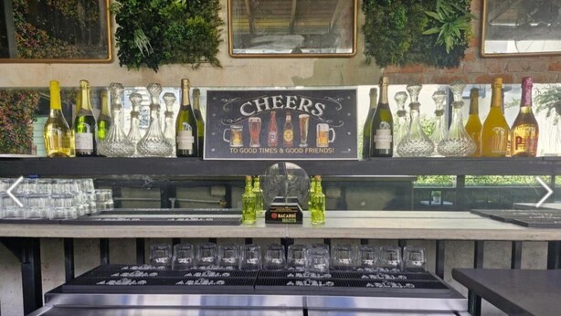 Zona de bar con un letrero que dice 'CHEERS TO GOOD TIMES & GOOD FRIENDS!' y una variedad de botellas y vasos.