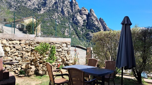 An outdoor patio with wicker furniture, a table, and an umbrella, set against a mountainous backdrop.