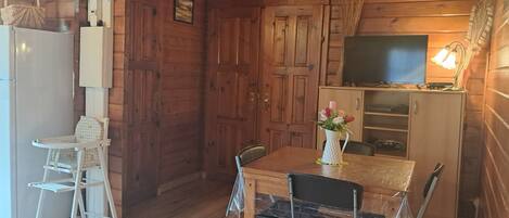 A wooden cabin interior with a dining table, chairs, a television, and a refrigerator.