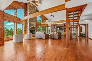 A spacious living area with wooden floors, a stone fireplace, and large windows offering a view of the outdoors. - Hill Country Retreat Near Villa Antonia - Two Homes, Pool, Lake Views & Private Trails (Jonestown)