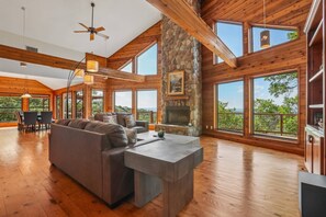 A spacious living room with a stone fireplace, wooden beams, large windows, and a ceiling fan. - Hill Country Retreat Near Villa Antonia - Two Homes, Pool, Lake Views & Private Trails (Jonestown)