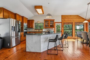 A modern kitchen with wooden cabinets, a stainless steel refrigerator, a marble island, and bar stools. - Hill Country Retreat Near Villa Antonia - Two Homes, Pool, Lake Views & Private Trails (Jonestown)