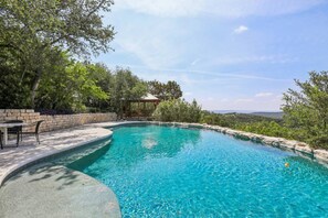 A swimming pool with a stone border, surrounded by trees and a stone wall. - Hill Country Retreat Near Villa Antonia - Two Homes, Pool, Lake Views & Private Trails (Jonestown)