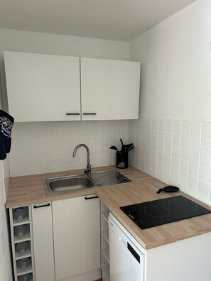 Et lille køkken med hvide skabe, en stålvask, en sort induktionskogetop og et opvaskestativ.