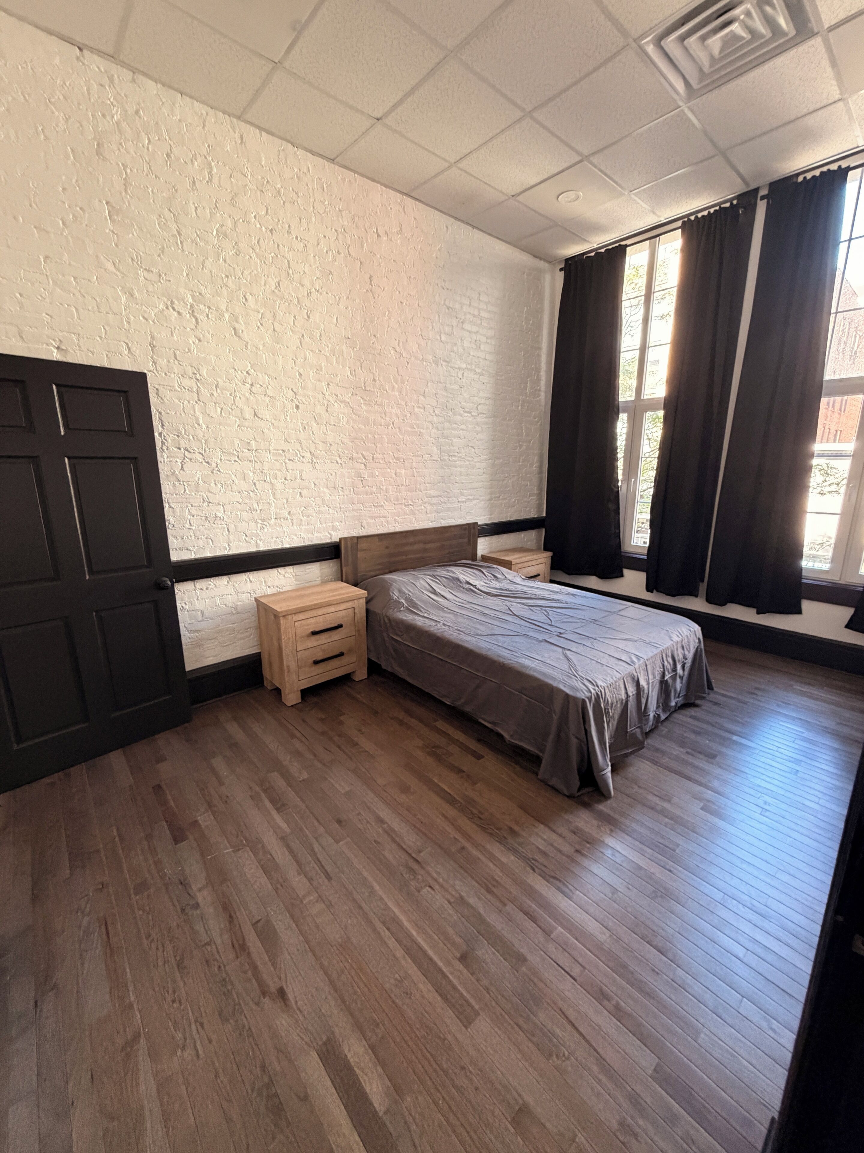 Ein Schlafzimmer mit Holzboden, einem Bett mit grauer Bettdecke, einem Nachttisch, einer schwarzen Tür und einem Fenster mit Vorhängen.