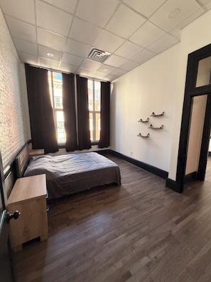A bedroom with a bed, a wooden desk, a window with curtains, a wall-mounted shelf, and a door.