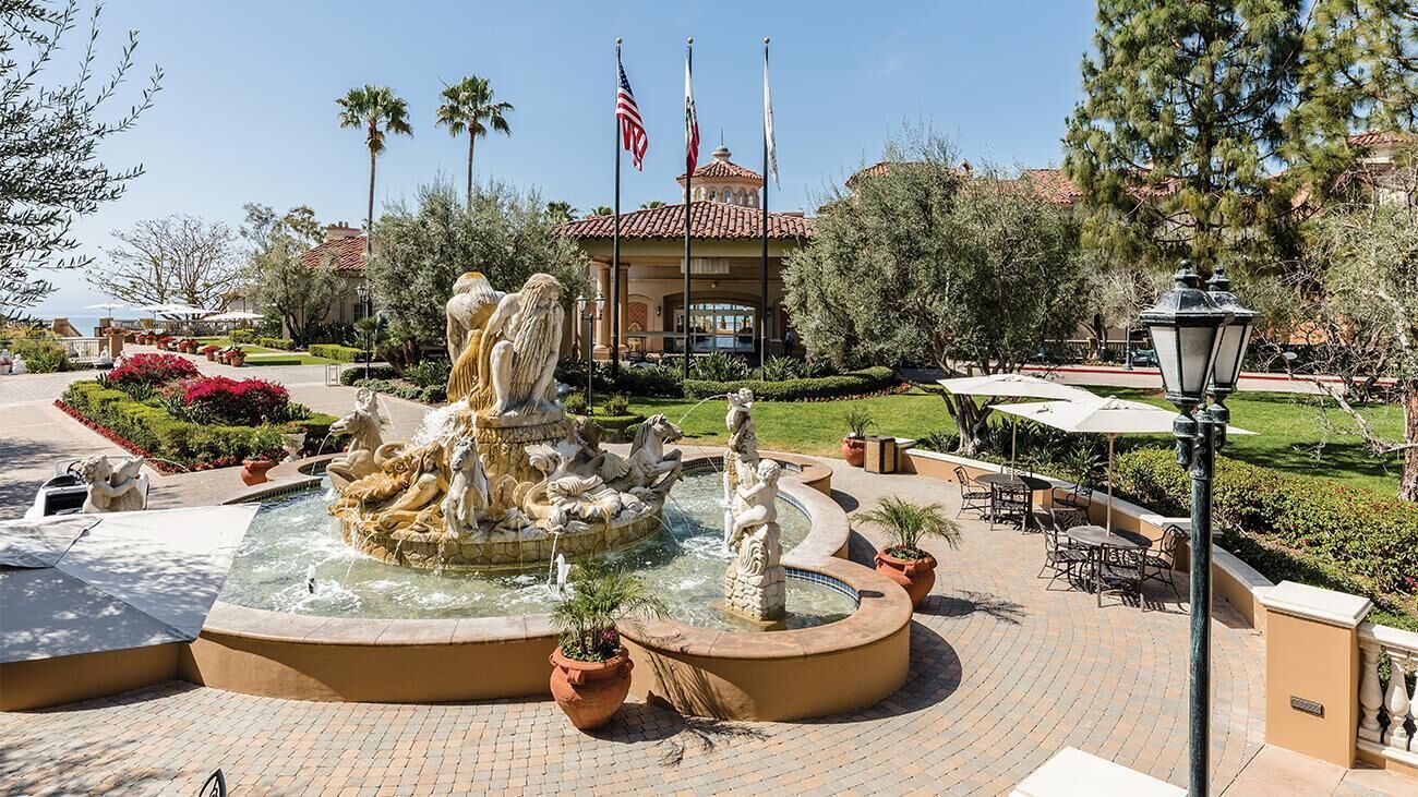 Una fuente con estatuas, un edificio con banderas y mesas al aire libre.