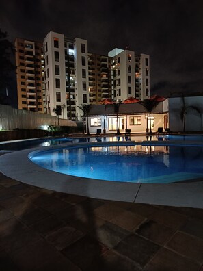Une piscine à bord incurvé, entourée d’une terrasse carrelée. Au fond, on aperçoit des immeubles résidentiels à plusieurs étages dotés de balcons.