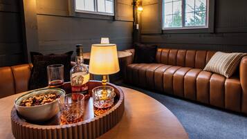 A cozy room with a leather couch, a round table with a lamp, a bowl of nuts, and a bottle of whiskey.