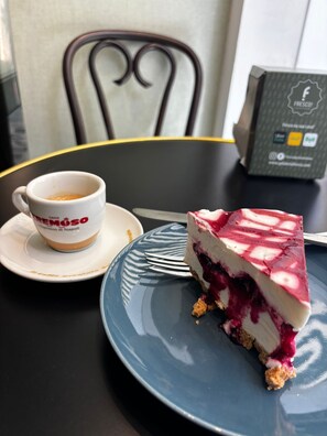 Ein Stück Käsekuchen mit Beeren auf einem blauen Teller, eine Kaffeetasse und ein Speisekartenständer auf einem Tisch.