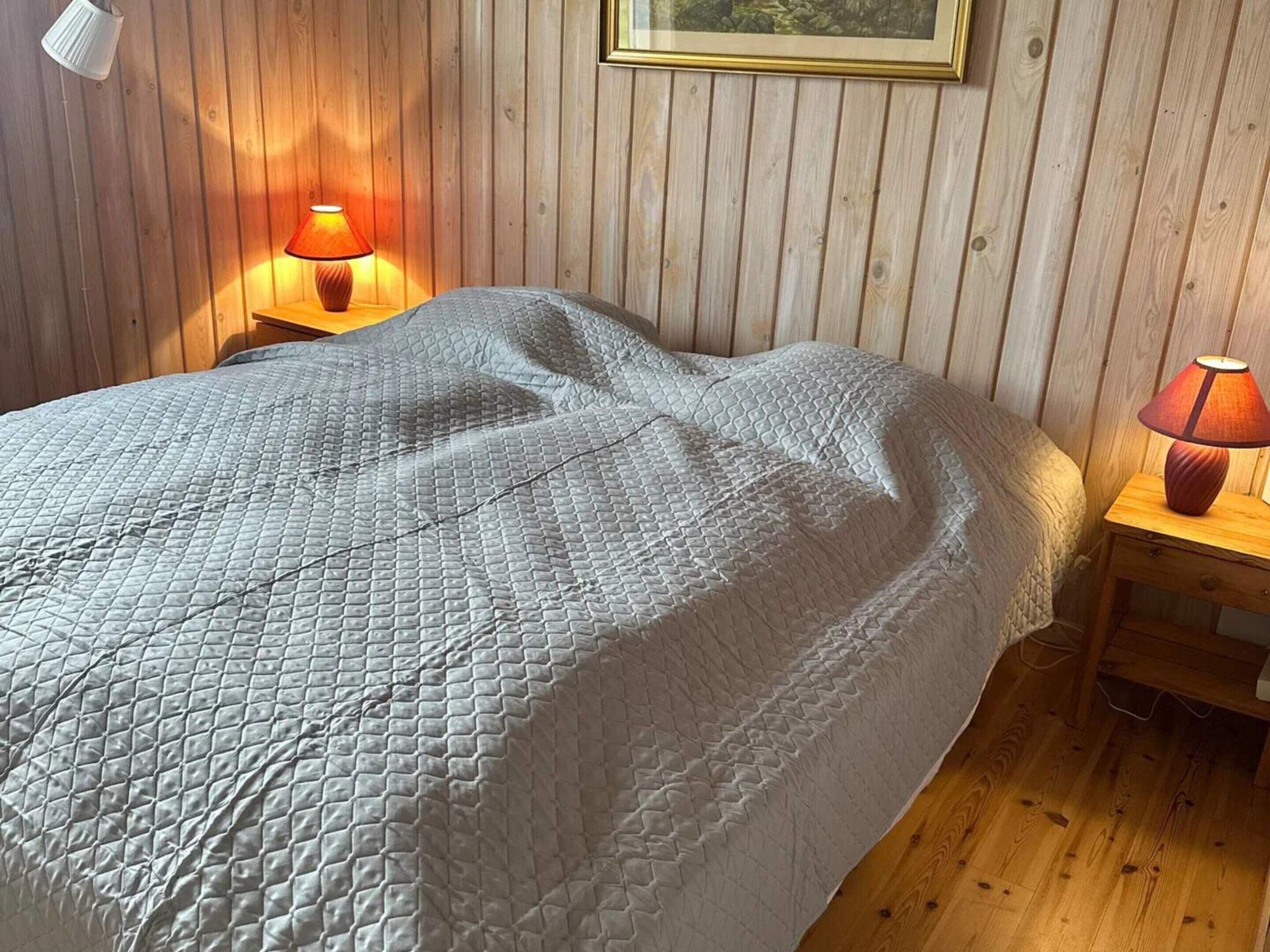 Een bed met een gewatteerde deken, twee nachtlampjes en een houten wand met een ingelijste foto.