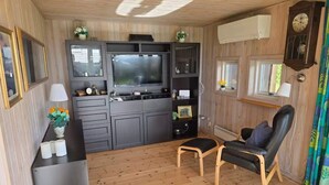 A compact living space with a kitchenette, a flat-screen TV, a wooden floor, a black leather chair, and a window with curtains.