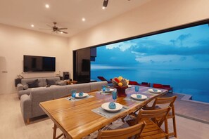 Ein modernes Wohnzimmer mit einem großen Esstisch, einer Couch und einem Fernseher. Durch ein großes Fenster bietet sich ein Blick auf den Ozean.
