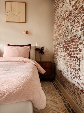 Un dormitorio con pared de ladrillo, una cama con ropa de cama rosa, una mesita de noche con un jarrón e iluminación de pared.
