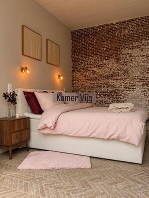 Un dormitorio con pared de ladrillo, una cama con colcha rosa, una mesita de noche con un jarrón y luces fijadas en la pared.