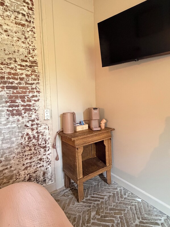 Una habitación con pared de ladrillo, una mesita de noche de madera con cafetera, un televisor montado en la pared y un suelo estampado.