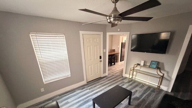 A living room with a ceiling fan, a TV mounted on the wall, a black leather couch, and a wooden floor. - Charming 3-bedroom house in brilliant Fort Worth with AC (Fort Worth)