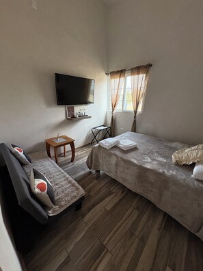 Una habitación pequeña y ordenada con cama, sofá, televisor montado en la pared, una mesita y una ventana con cortinas.