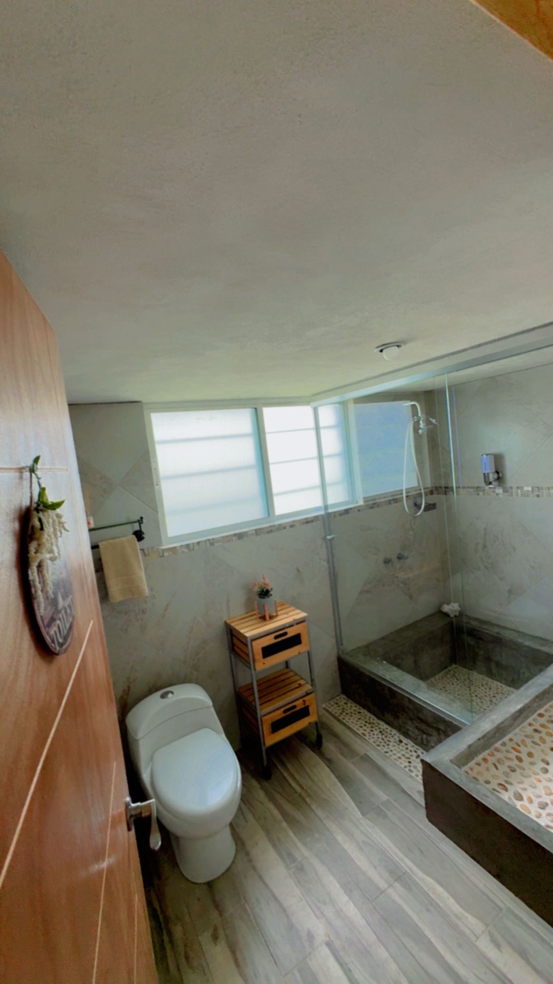 Een badkamer met een toilet, een houten kast, een glazen douchecabine en een raam.