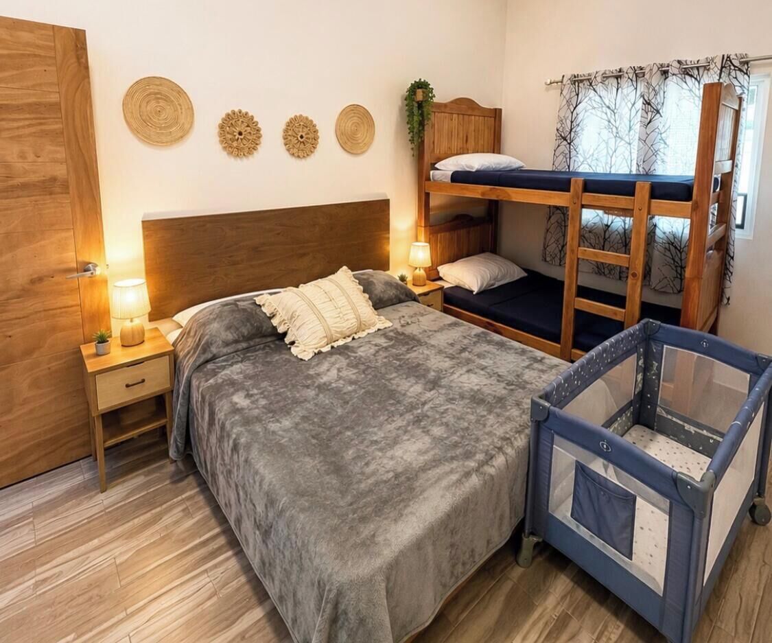 A bedroom with a bunk bed, a single bed, a nightstand with a lamp, and a baby playpen.