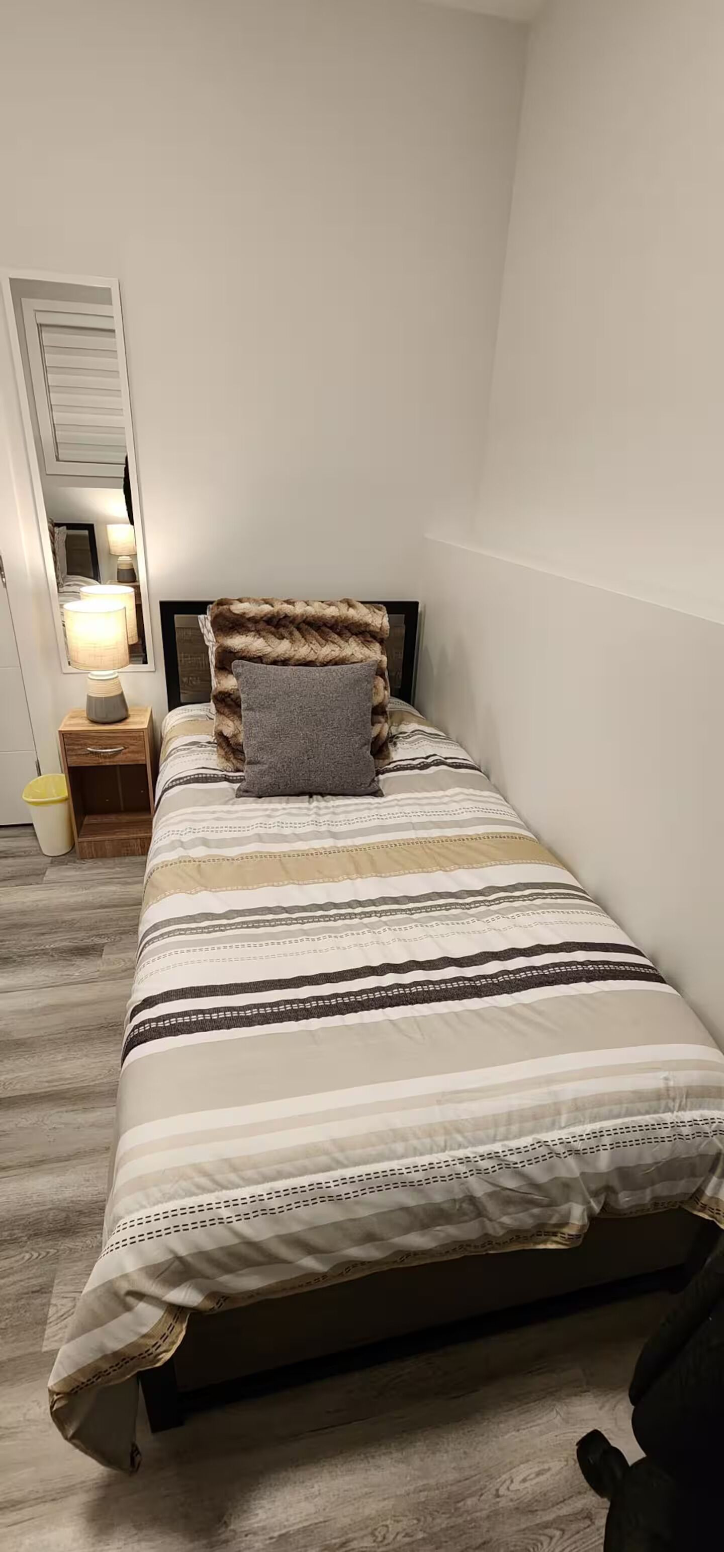 Una cama individual con edredón a rayas, una lámpara de noche, un espejo y un cubo de basura.