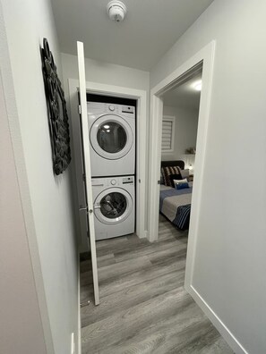 Ett tvättrum med två staplade tvättmaskiner och en hall som leder till ett sovrum med en säng och ett nattduksbord.