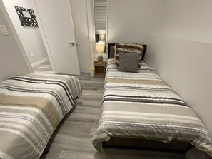 Et soveværelse med to enkeltsenge, et natbord med lampe, og en dør der fører til et andet rum.