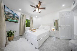 A modern bedroom with a bed, a ceiling fan, a bedside table, a nightstand, a large window, a wall-mounted TV, and a door. - Adorable 2-bedroom house next to Hard Rock Casino (Fort Lauderdale)