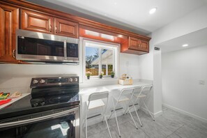A kitchen with wooden cabinets, a stainless steel microwave, a black stove, and a window with a view of a house and trees. - Adorable 2-bedroom house next to Hard Rock Casino (Fort Lauderdale)