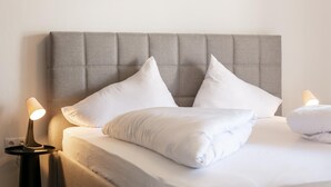 Ein ordentlich bezogenes Bett mit weißer Bettwäsche und Kissen, ein grau gepolsterter Kopfteil und zwei Nachttischlampen auf einem Nachttisch.