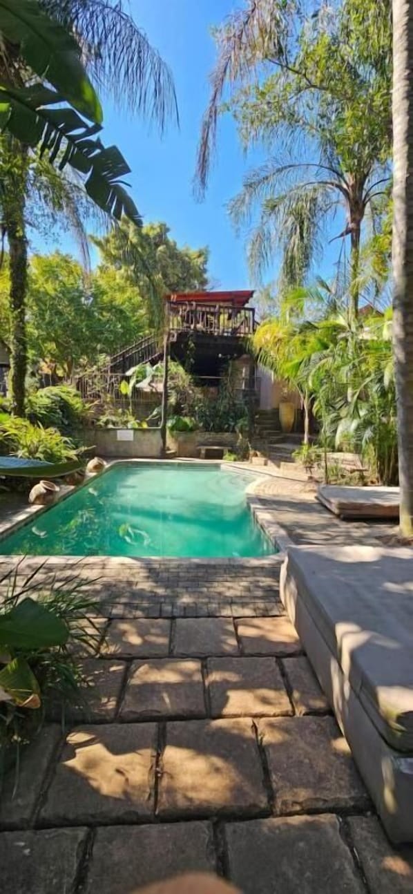 A swimming pool surrounded by stone steps and lush greenery, with a wooden deck and a building in the background. - Old Vic Travellers Inn (Mbombela)