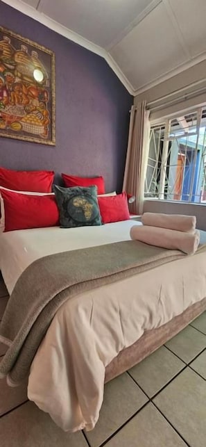 A bed with red and green pillows, a beige blanket, and a window with curtains. - Old Vic Travellers Inn (Mbombela)