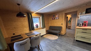 A cozy room with a wooden ceiling, a dining table with chairs, a bench, a radiator, a window with curtains, a kitchen area with a microwave and a coffee maker, and a rug on the floor. - LostGarden Öblarn Top 1 (Öblarn)