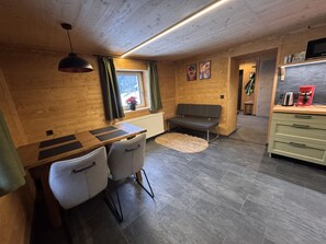 A cozy room with a wooden ceiling, a dining table with chairs, a bench, a radiator, a window with curtains, a kitchen area with a microwave and a coffee maker, and a rug on the floor. - LostGarden Öblarn Top 1 (Öblarn)
