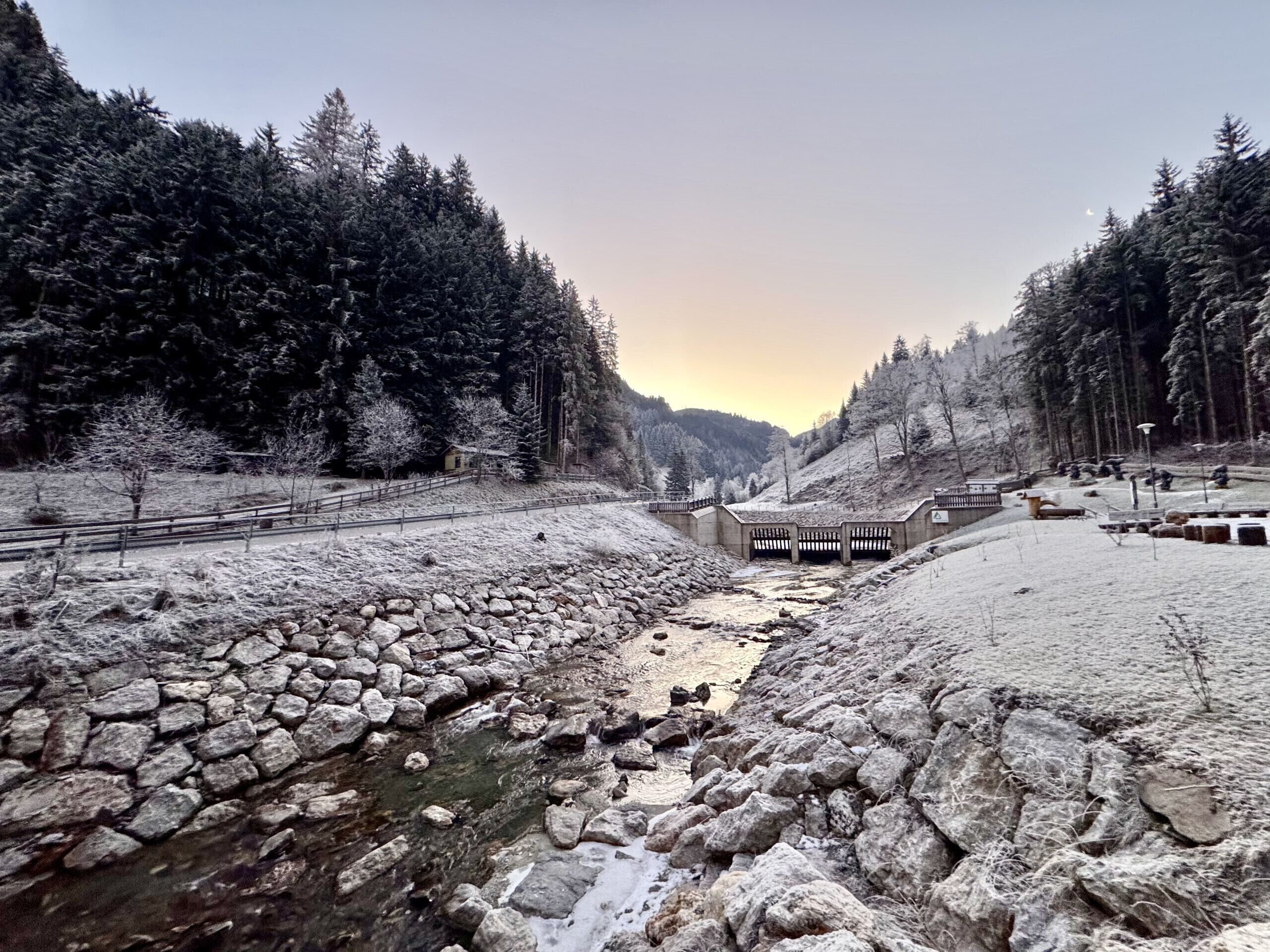 Un río con lecho pedregoso, rodeado de árboles cubiertos de nieve y un puente de madera.
