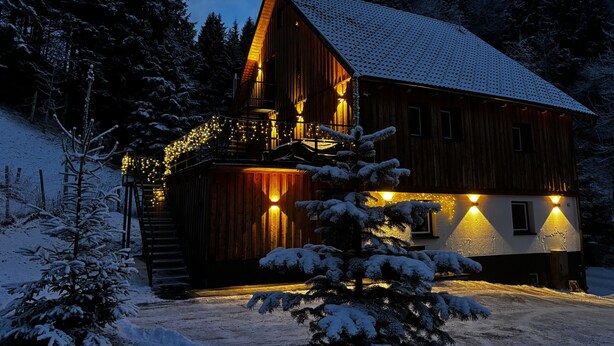 A wooden cabin with a sloped roof, surrounded by snow-covered trees, and illuminated by warm lights. - LostGarden Öblarn Top 1 (Öblarn)