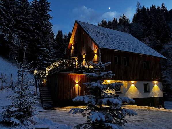 A wooden cabin with a sloped roof, surrounded by snow-covered trees, and illuminated by warm lights. - LostGarden Öblarn Top 1 (Öblarn)