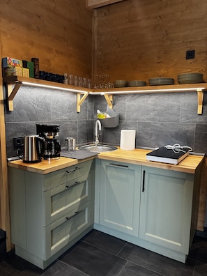 A kitchen with a wooden countertop, a coffee maker, a sink, and a shelf with plates and glasses. - LostGarden Öblarn Top 1 (Öblarn)