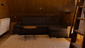 A cozy loft bedroom with a wooden ceiling, a black sectional sofa, a small round table, and a ladder. - LostGarden Öblarn Top 3 (Öblarn)