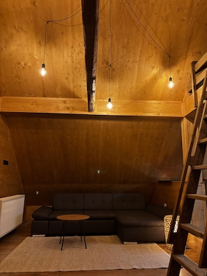 Ein gemütliches Schlafzimmer im Loft-Stil mit Holzbalkendecke, einem schwarzen Ecksofa, einem kleinen runden Tisch und einer Leitern.