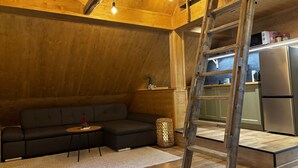 A loft-style room with a wooden ceiling, a sofa, a small table, a ladder, and a kitchen area with a refrigerator. - LostGarden Öblarn Top 3 (Öblarn)