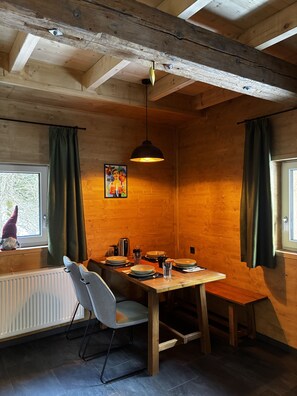 A wooden interior with a dining table set for four, a chair, a hanging light fixture, and a window with curtains. - LostGarden Öblarn Top 2 (Öblarn)