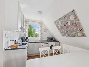 A modern kitchen with white cabinetry, a window with curtains, a microwave, a washing machine, and a dining area with a white table and chairs. - Cozy holiday home by the Baltic Sea for groups (Bastorf)