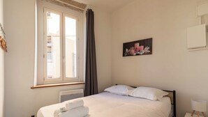 Une chambre à coucher avec un lit, une fenêtre avec des rideaux, un radiateur, un tableau accroché au mur et une table de chevet avec une lampe.