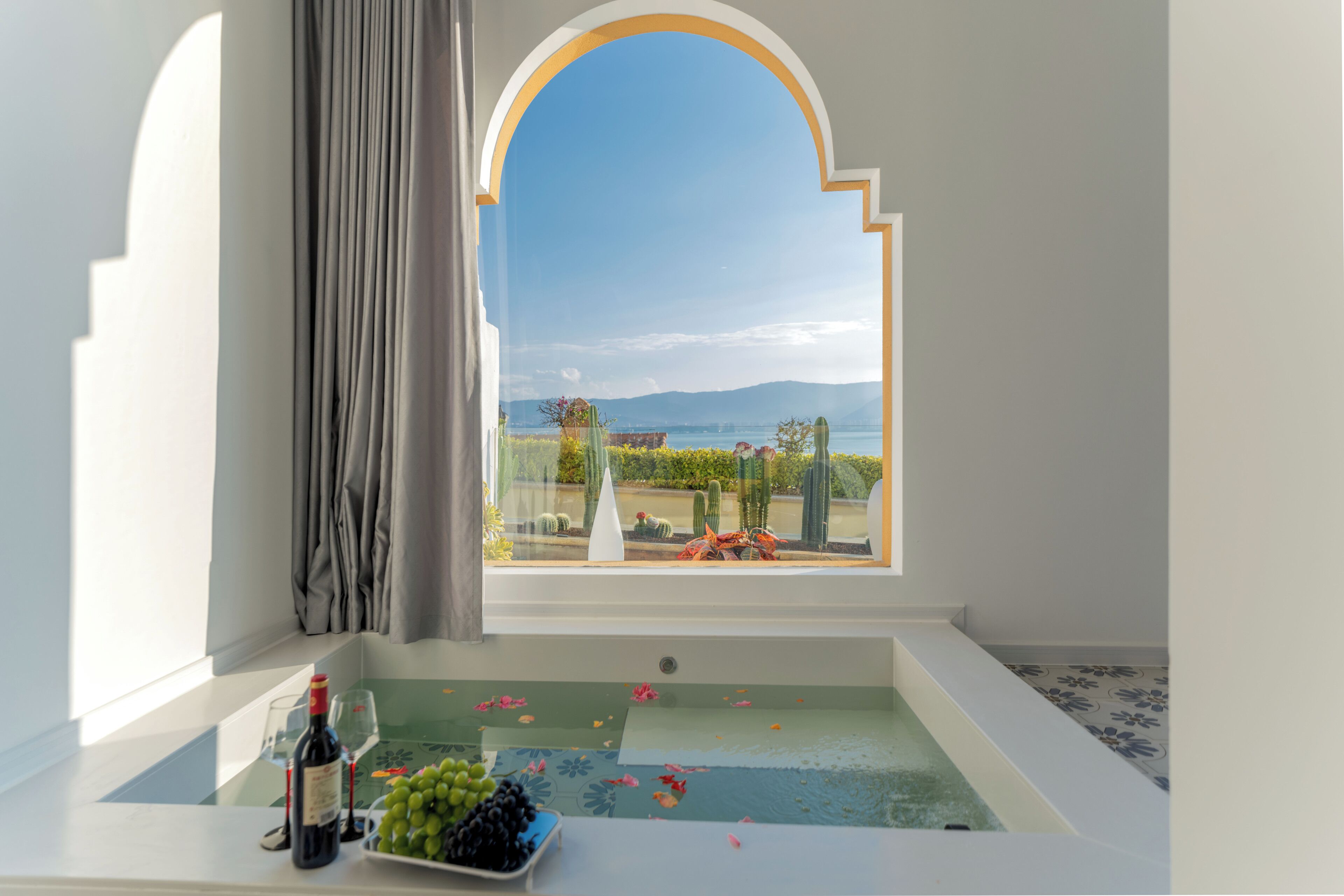A room with a bathtub, a window with a view of a garden and mountains, and a table with a bottle and grapes.