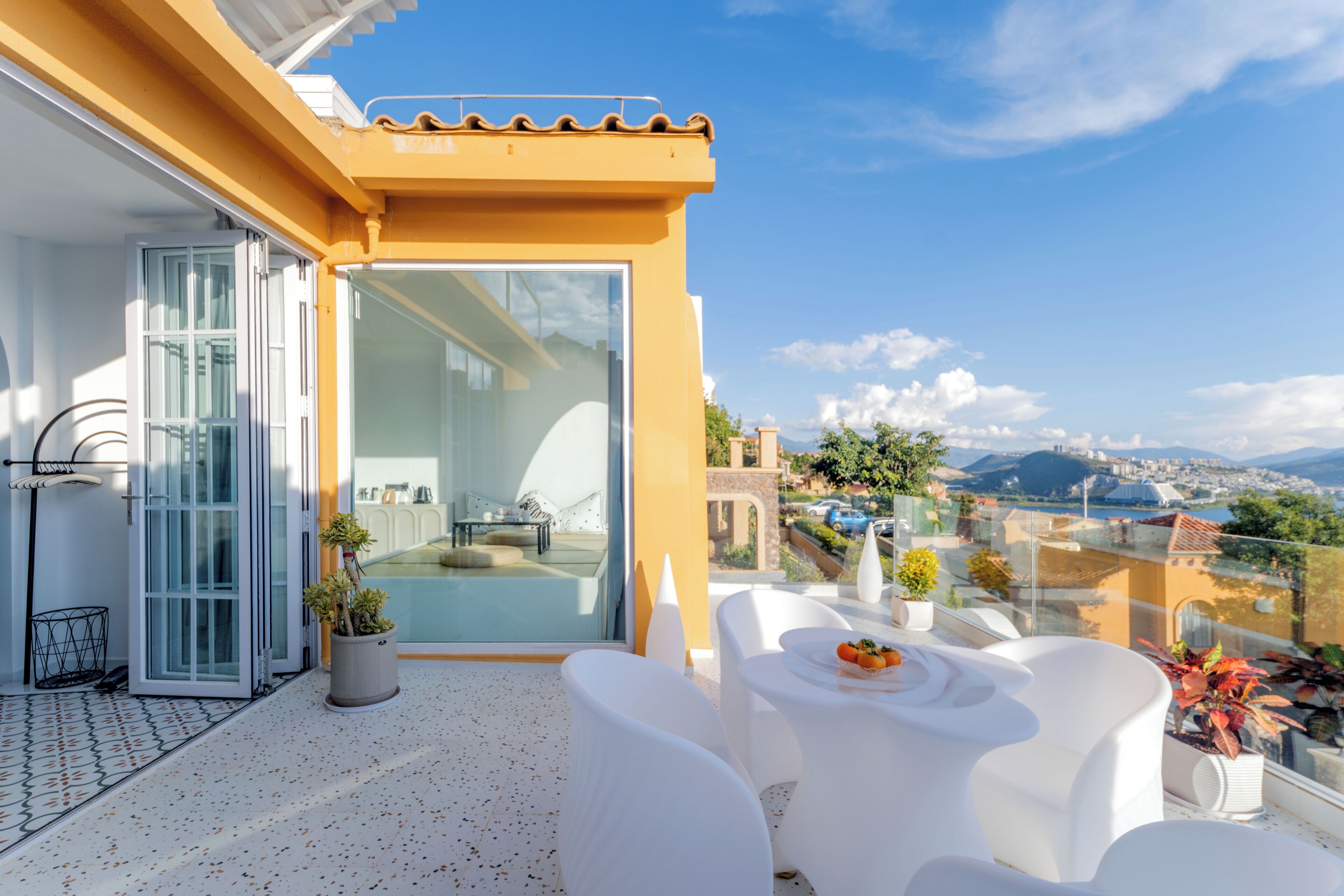 Un balcón con mobiliario blanco, una puerta de cristal y vistas a un paisaje urbano.