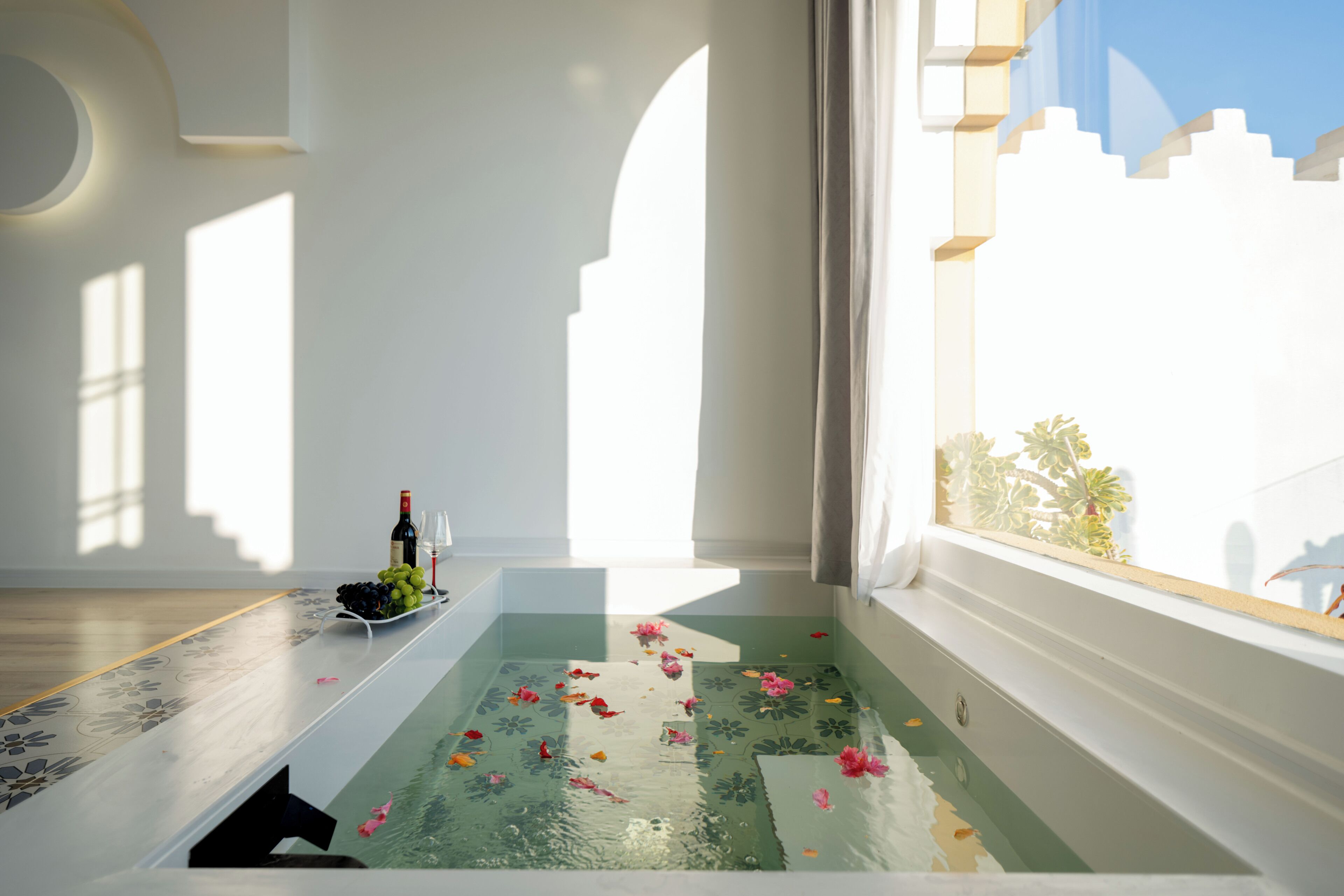 A room with a bathtub filled with water and rose petals, a bottle of wine, and a glass on a table.