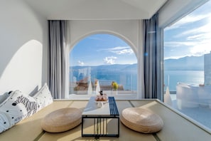 Una stanza moderna con un'ampia finestra ad arco, un divano con cuscini decorati, un tavolino con oggetti ornamentali e vista sul mare e sulle montagne.