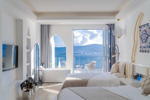 Habitación de hotel con una cama grande, una cama más pequeña, un balcón y vistas al mar, además de un comedor.